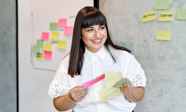 Floria Moghimi hält Post its in der Hand und steht vor einer Wand auf der Post its kleben. In dem Workshop geht es um Diversity-Strategie. Sie blickt nach rechts aus dem Bild.